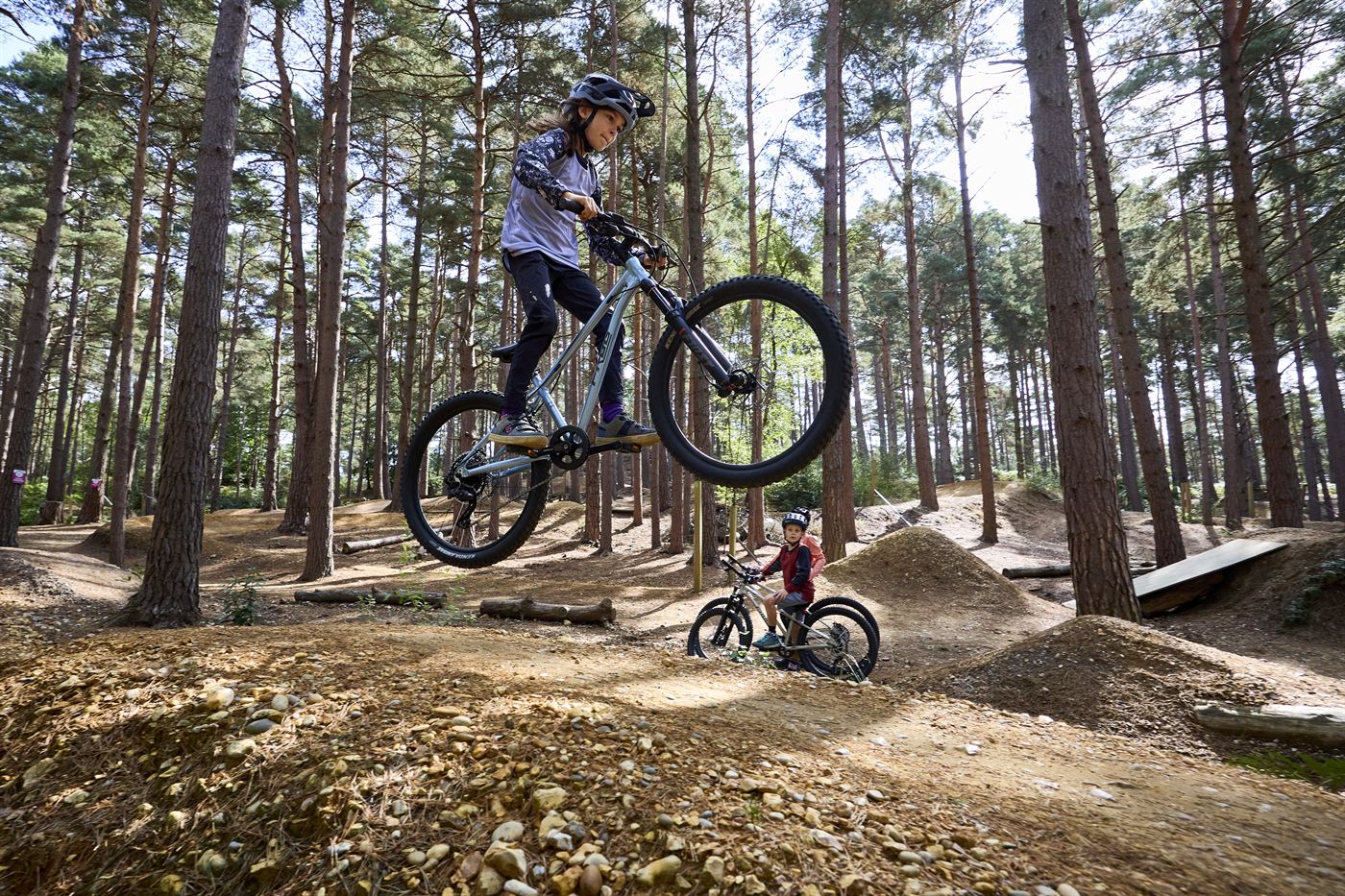 A child jumping a Frog mountain bike on a woodland trail while another rider watches